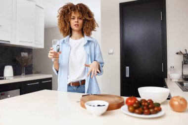 Sıcak bir mutfak ortamında elinde şarap kadehiyle meyve ve sebzelerin önünde duran bir kadın.