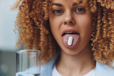 Woman with curly hair sticking out tongue holding a pill, concept of playful defiance against medication clipart