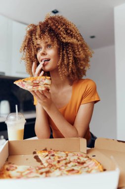 Genç bir kadın pizza kutusunun önünde kıvırcık saçlı lezzetli bir pizzanın tadını çıkarıyor.