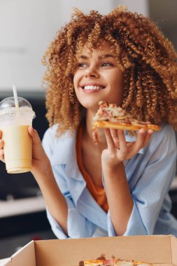 Kıvırcık saçlı kadın bir dilim pizza ve smoothie 'yi masanın üstünde pizza kutusunun önünde tutuyor.
