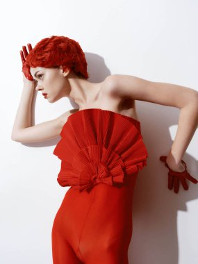 Kendine güvenen kırmızı elbiseli, elleri kalçasında ve rahat ifadesiyle beyaz bir duvara poz veren, kendine güvenen bir kadın.