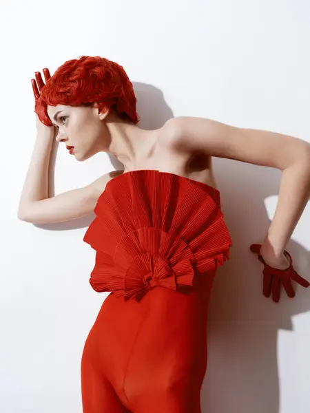 Confident woman in a vibrant red dress posing against a clean white wall with hands on hips and relaxed expression