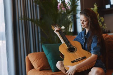 Kanepede oturan genç bir kadın pencerenin önünde akustik gitar çalıyor ve dingin bir müzik ortamı yaratıyor.