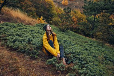 Sonbahar güzeli sarı ceketli genç bir kadın ormanda oturuyor tepeler ve ağaçlarla arka planda