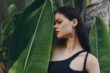 Siyah mayo giymiş genç bir kadın tropikal plaj tatil beldesindeki yemyeşil yapraklarla palmiye ağacına yaslanıyor.