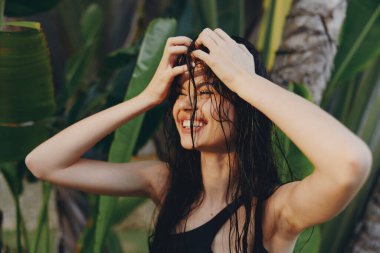 Güneşli bir günde, elleri başının üstünde palmiye ağacının önünde duran, ıslak saçlı, tropik tatil kadını.