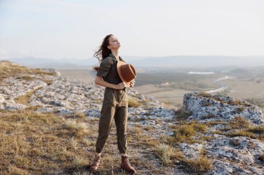 Tepede durmuş şapkalı bir kadın gökyüzüne bakıyor doğanın güzelliğini ve seyahat ilhamını düşünüyor.