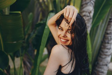 Uzun siyah saçlı egzotik güzellik. Palmiye ağaçlarının önünde elleri kafasında poz veriyor.