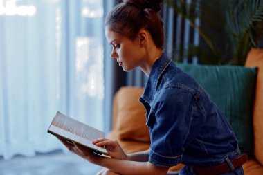 Kadın evde oturup kitap okuyor. Kanepede, pencerenin önünde doğal ışıkta.