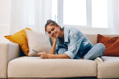 Gülümseyen kadın cep telefonunu tutuyor ve rahat oturma odasında koltukta dinleniyor.