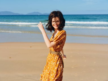 Turuncu elbiseli mutlu kadın kumsalda güneşli bir günün tadını çıkarıyor elleri havada.