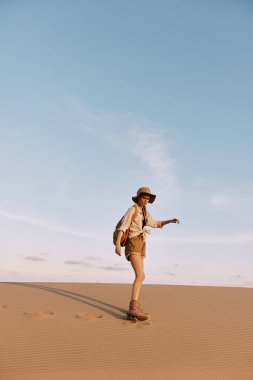 Şapkalı kadın kumlu çölde yürüyor açık mavi gökyüzünün altında manzaralı bir yerde.