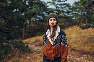 Şık ceketli ve şapkalı şık bir genç kadın sakin ormanda manzaralı bir toprak yolda yürüyor.