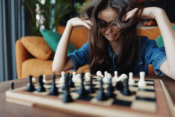 Stressed woman playing chess at table in front of couch with head in hands