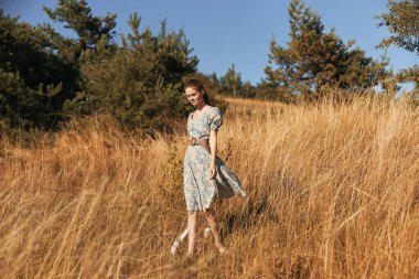 Çiçekli ağaçlar ve uzun otlarla çevrili sakin bir tarlada dikilen elbiseli bir kadın.