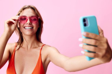 Canlı ve neşeli, renkli mayo giymiş genç bir kadın pembe arka planda telefonla selfie çekiyor.