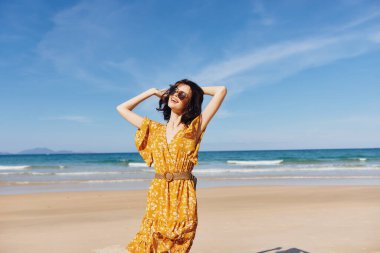 Parlak sarı elbiseli bir kadın elleri kumlu sahilde duruyor ve arkasında okyanus dalgaları var.