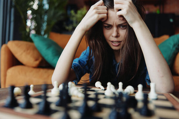 A thoughtful woman sitting on a couch playing chess game with hands on head in living room