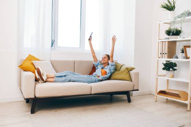Cozy Sofa 'da akıllı telefon tutan neşeli kadın çevrimiçi oyunların tadını çıkarıyor ve evinin rahatlığında sohbet ediyor.