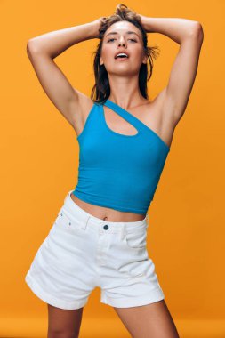 Şık mavi şortlu ve beyaz şortlu kendine güvenen genç bir kadın neşeli yaz moda kavramında canlı turuncu arka planına karşı elleri başının üstünde oyuncu pozlar veriyor.