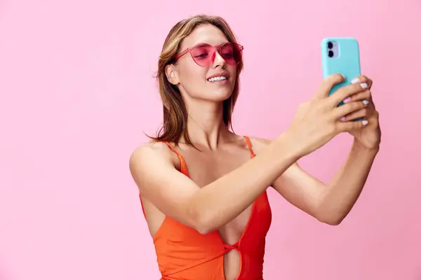 stock image Vibrant woman in bright orange swimsuit and sunglasses captures perfect beach moment with selfie on cell phone