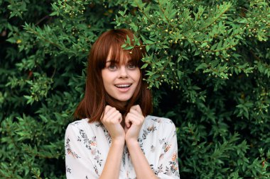 Canlı kızıl saçlı kadın canlı yapraklarla yemyeşil çalıların yanında zarif bir şekilde duruyor.