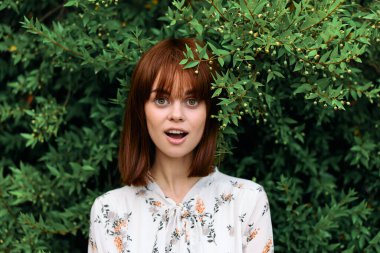 Canlı kızıl saçlı kadın, gür yeşil yapraklı çalıların önünde zarifçe poz veriyor.