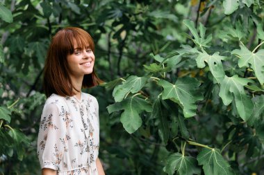 Parlak yeşil parkta ağaçlar ve yapraklarla poz veren canlı kızıl saçlı kadın.