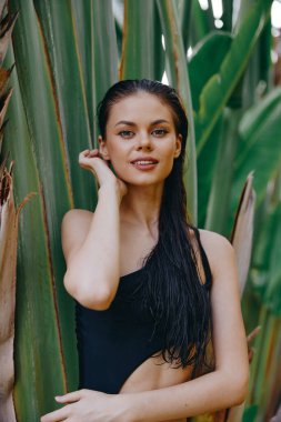 Plajdaki egzotik güzellik siyah mayo giymiş çekici genç kadın tropik palmiye ağaçları ve yemyeşil bitkiler tarafından poz veriyor.
