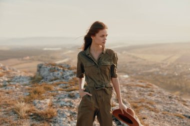 Yeşil tulumlu kadın, seyahat ve macera konsepti için şık pozlarda şapkalı tepenin üstünde duruyor.
