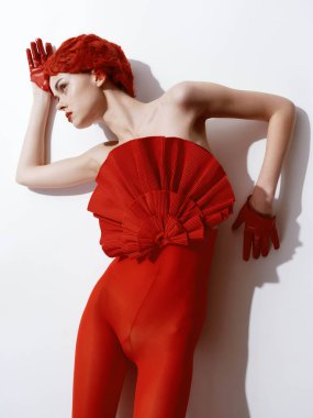 Minimalist moda çekiminde beyaz duvara poz veren kırmızı elbiseli güzel kızıl saçlı kadın.