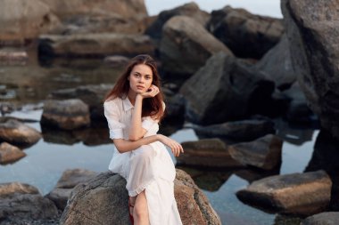 Huzurlu beyaz elbiseli kadın sakin sularda, elleri kayalık kıyılarda çene üzerinde tefekkür ediyor.