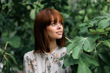 Canlı kızıl saçlı bir kadın dingin bir parkta yemyeşil yeşilliklerin önünde zarif bir şekilde duruyor.