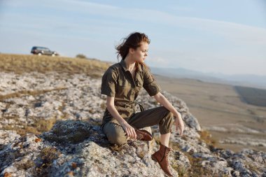 Dağın tepesinde oturan yeşil gömlekli ve kahverengi çizmeli kadın doğanın güzelliği içinde doğa ve maceranın tadını çıkarıyor.