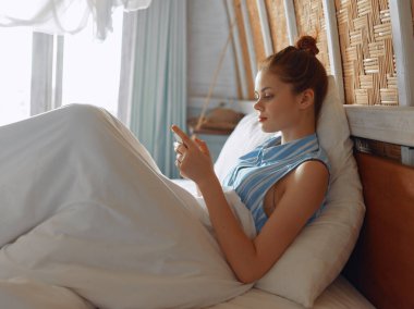 Yatakta rahat beyaz gömlek ve mavi pijama pantolon giyen huzurlu bir kadın telefonunu rahat bir ifadeyle kontrol ediyor..