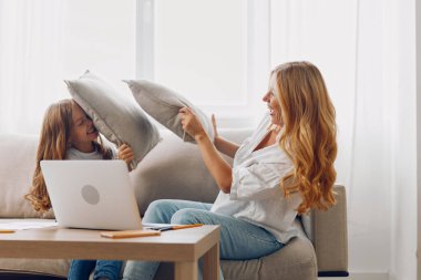 Anne ve kızı kanepede yastıklarla kaynaşıyor, rahat bir aile anı için dizüstü bilgisayarlarını paylaşıyorlar.