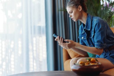 Ev Kadınları 'nda dinleniyor. Bir kase meyve ve cep telefonuyla koltukta oturuyor.