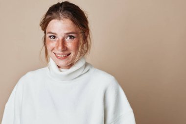 Beyaz balıkçı yaka kazaklı güzel bir kadın bej duvarın önünde duruyor.