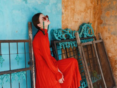 Çarpıcı kırmızı elbiseli kültürlü bir kadın canlı mavi duvarın ve pencerenin önünde zarif bir şekilde duruyor..