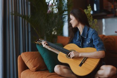 Pencerenin önündeki koltukta akustik gitar çalan genç bir kadın.