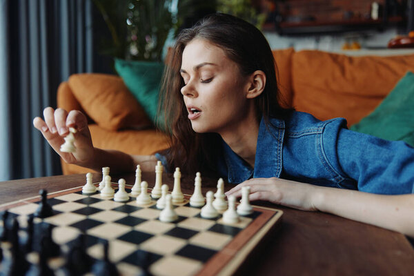 Woman playing chess game on table in front of couch with lamp, book and plant in background