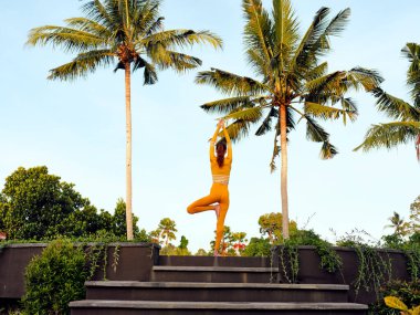 Sarı elbiseli kadın bereketli tropikal ortamda yoga yapıyor. Palmiye ağaçları ve basamaklarla çevrili. Huzurlu ve rahatlatıcı bir atmosfer yaratıyor..