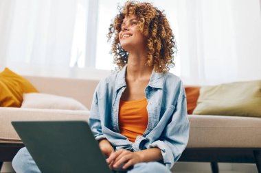 Modern oturma odasındaki rahat koltukta dizüstü bilgisayarla çalışan, kulaklıkları ve telefonu olan gülümseyen bir kadın.