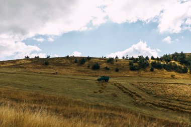 Tarlada traktörü olan kırsal alan, yuvarlanan tepeler, ve gökyüzünde dramatik bulutlar.