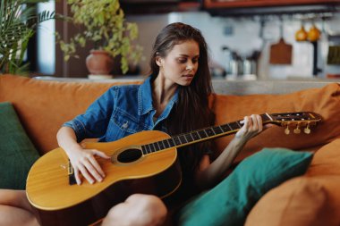 Oturma odasındaki kanepede akustik gitar çalan genç bir kadın rahat bir atmosferde yakalandı.