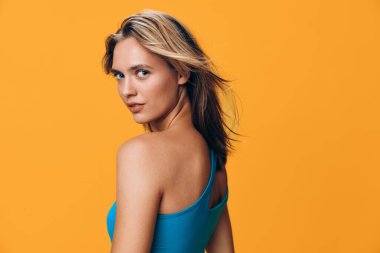 Mavi kolsuz tişörtlü vahşi ve kendinden emin bir kadın canlı turuncu bir arka plana karşı güçlü bir poz veriyor.