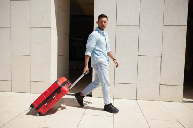 Açık mavi elbiseli, kırmızı bavulu çeken, modern bir mimari binanın önünden geçen seyahat ya da iş gezisi öneren kendine güvenen genç bir adam..