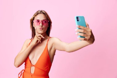 Turuncu mayo giymiş canlı bir kadın yumuşak pembe arka plan önünde selfie çekiyor.