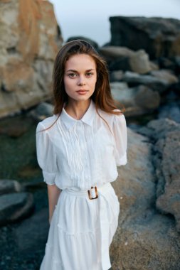 Okyanusa bakan kayalık kıyı şeridinde duran beyaz elbiseli sakin genç kadın.