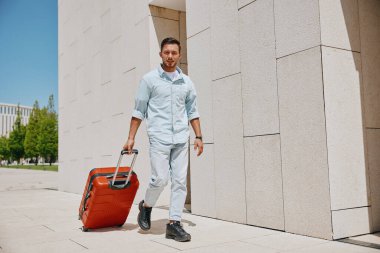 Kırmızı bavulu çeken, parlak, güneşli bir arka planı olan çağdaş bir binanın yanında yürüyen sıradan bir adam..